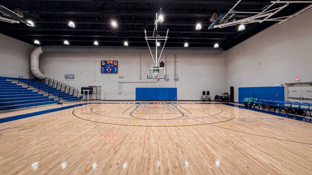 An empty indoor basketball court with polished wooden flooring, blue bleachers and wall padding.